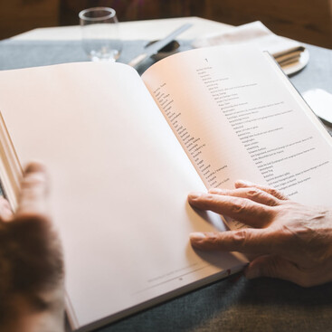 Nahaufnahme einer geöffneten Buchseite mit einer alphabetisch geordneten Liste von Dialektwörtern und deren Übersetzungen ins Hochdeutsche, gehalten von zwei Händen auf einem gedeckten Tisch.