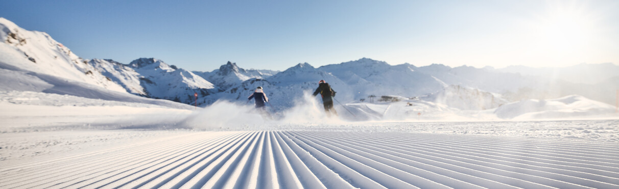 Zwei Skifahrer:innen gleiten bei strahlendem Sonnenschein über eine frisch präparierte Skipiste inmitten der verschneiten Berge von St. Anton am Arlberg. Die klaren Linien im Schnee betonen die perfekte Pistenqualität. Der Hintergrund zeigt das beeindruckende Alpenpanorama unter wolkenlosem Himmel.