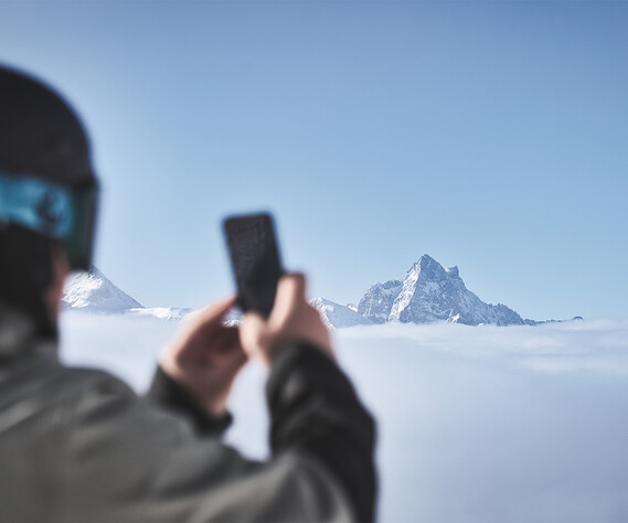 Ein:e Wintersportler:in hält ein Smartphone und fotografiert die markanten Gipfel der Arlberger Alpen, die aus einem dichten Wolkenmeer hervorragen. Im Vordergrund verschwommen sichtbar sind Helm und Skibrille, die den Fokus auf die majestätische Berglandschaft lenken.
