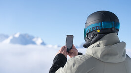 Skifahrer mit grauer SKijacke und schwarzen Helm macht ein Bild von der verschneiten Winterlandschaft in St. Anton am Arlberg