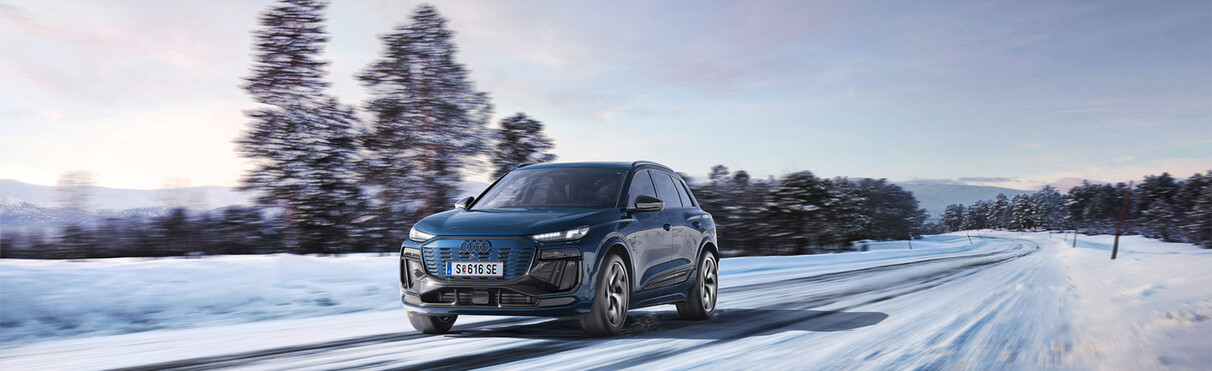 Dark grey Audi Q6 e-tron drives through a snowy scene, the road is only partially covered with snow.