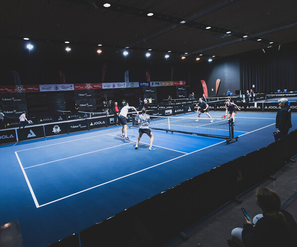 Zwei Teams spielen Pickleball auf einem beleuchteten Indoor-Spielfeld mit blauen Bodenlinien. Im Vordergrund ist ein dynamisches Spiel im Gange, während Zuschauer am Rand des Feldes sitzen.