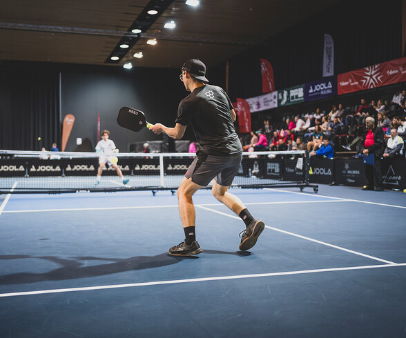 Ein junger Mann in Sportkleidung schlägt einen Pickleball auf einem Hallenspielfeld, im Hintergrund ist das Publikum auf den Tribünen sichtbar.