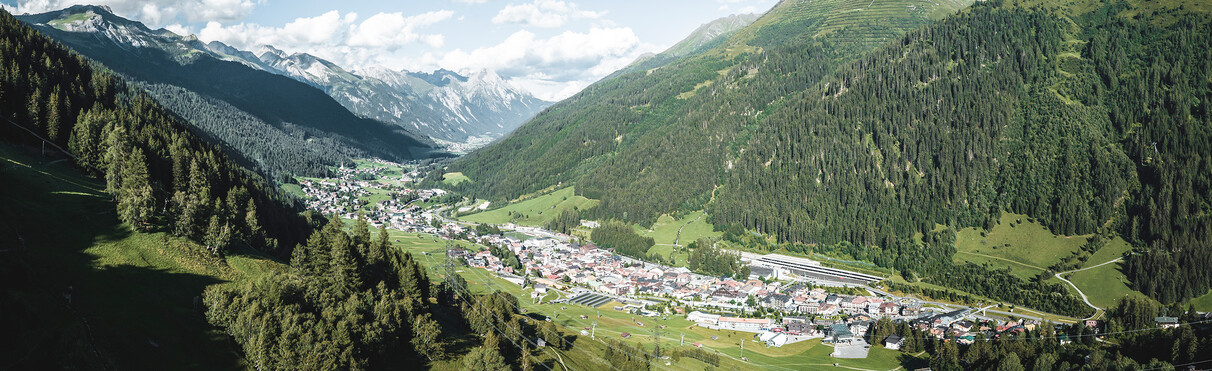 Wide view over St. Anton am Arlberg with dense forests, alpine peaks and the scenic village nestled in a green valley on a sunny day.