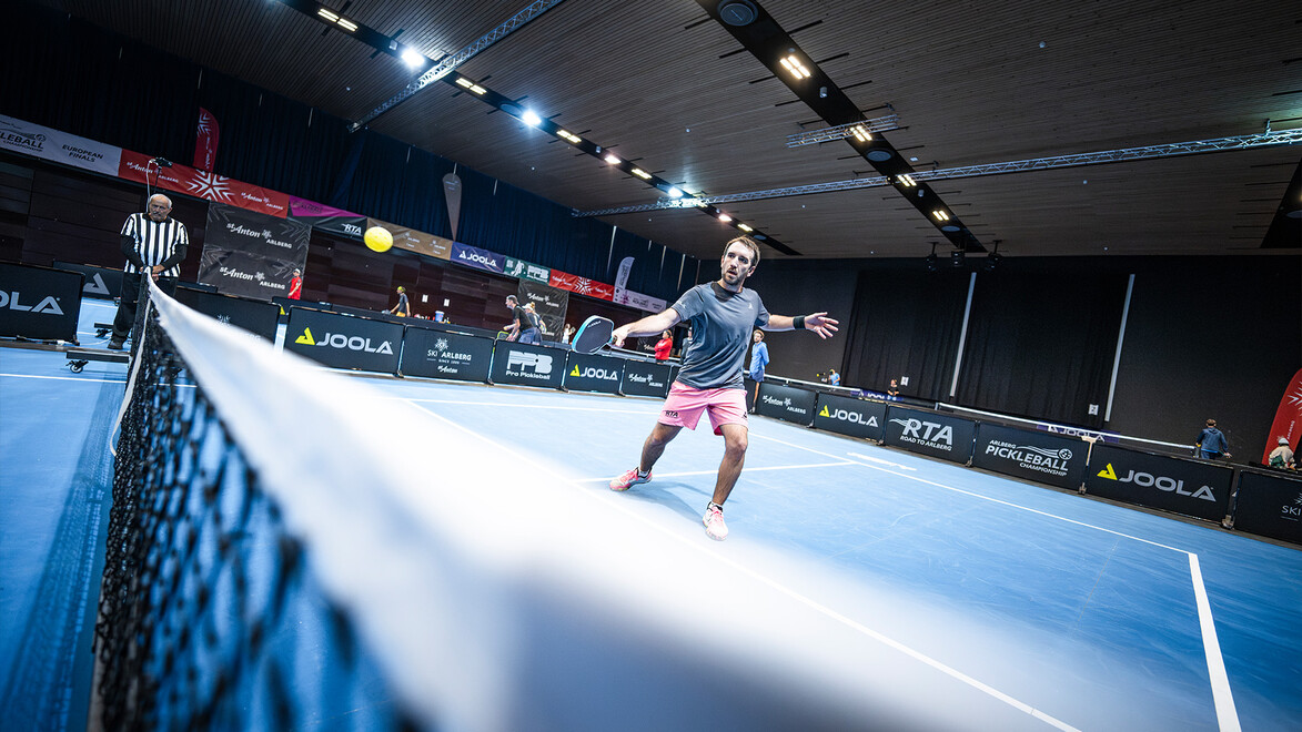 Ein Spieler in Aktion beim Aufschlag während eines Pickleball-Matches auf einem blau beleuchteten Hallenplatz / A player serves during a pickleball match on a blue-lit indoor court