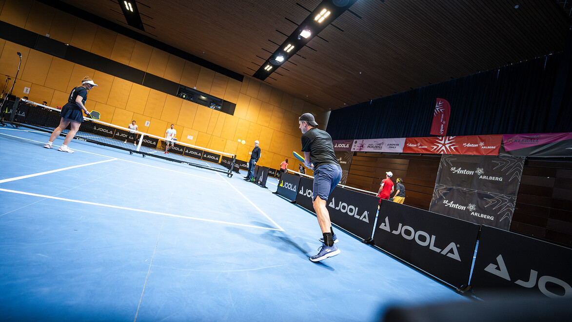 Zwei Spieler liefern sich ein schnelles Duell auf einem Hallenplatz mit Sponsorenbannern im Hintergrund / Two players in a fast rally on an indoor court with sponsor banners in the background