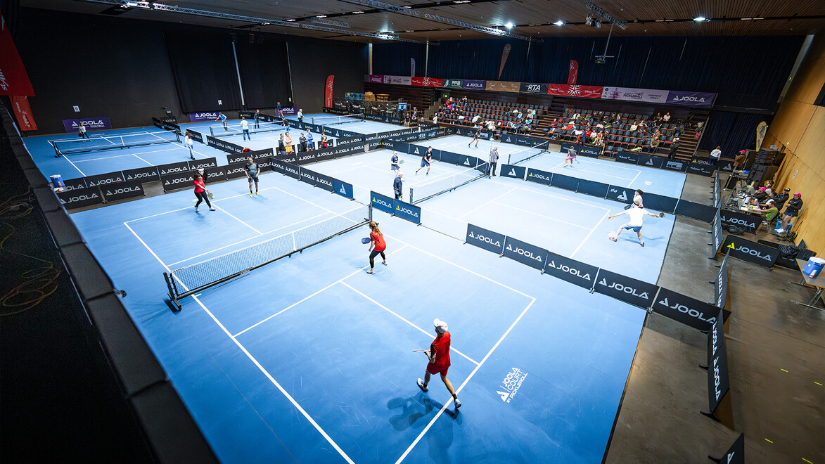 Blick auf mehrere Pickleball-Spielfelder in einer Sporthalle mit aktivem Spielbetrieb und Zuschauerbereich / View of several pickleball courts in a sports hall with active gameplay and spectator area