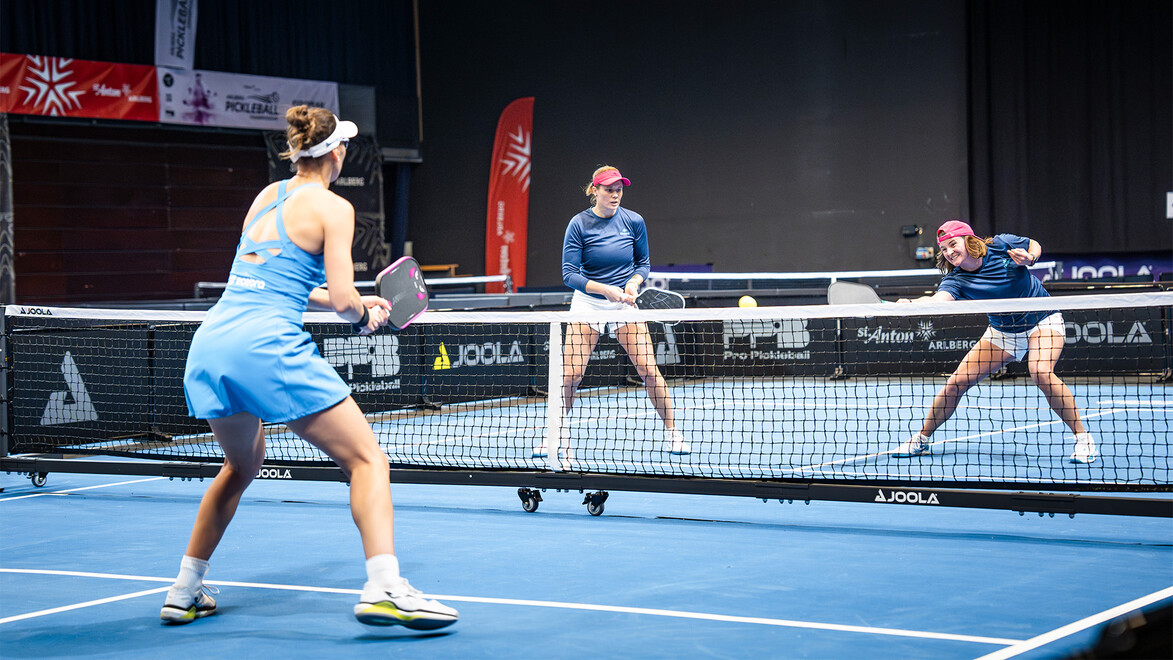 Drei Frauen spielen ein spannendes Pickleball-Doppelmatch in einer modernen Halle mit blauen Spielfeldern / Three women are playing a thrilling pickleball doubles match in a modern indoor venue with blue courts