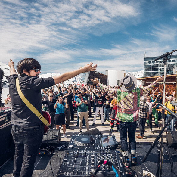  Zwei Musiker auf einer Outdoor-Bühne heizen der Menge ein, während zahlreiche Gäste vor verschneiter Bergkulisse feiern.