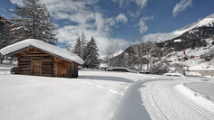 Eine gespurte Langlaufloipe führt an einer schneebedeckten Hütte vor Bergkulisse vorbei.