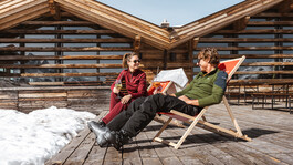 Zwei Personen entspannen auf Sonnenliegen vor einer Berghütte mit Schnee und Winterstimmung.