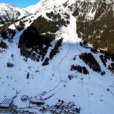 Luftaufnahme einer verschneiten Berglandschaft mit der markierten Weltcup-Skipiste und Zielbereich in St. Anton am Arlberg, eingerahmt von dichten Wäldern und Gebäuden im Tal.