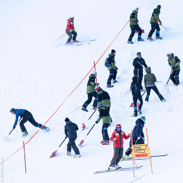 Mehrere Personen mit Schaufeln und Skiausrüstung arbeiten am präparieren einer Skipiste vor einem Weltcuprennen, während ein Sicherheitsläufer vorbeifährt.