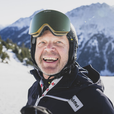 Nahaufnahme von Rudi Lapper mit Skibrille und Helm vor verschneiten Bergen, der eine Jacke mit dem Aufnäher „Schöffel“ trägt.