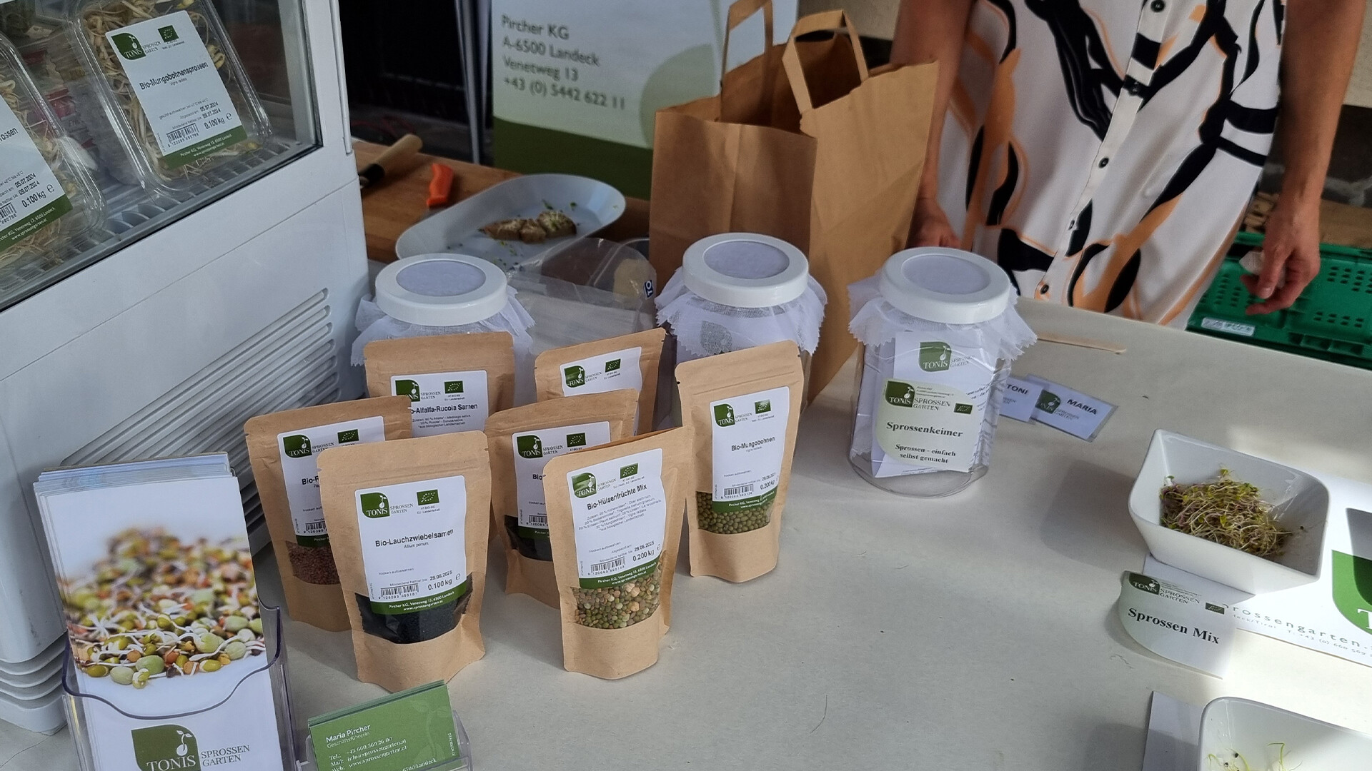 Produkte aus dem Sprossengarten wie Samen und Keimlinge auf einem hellen Verkaufstisch am Markt präsentiert. / Products from the sprout garden like seeds and seedlings presented on a bright sales table at the market.