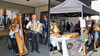 Links Musikanten mit Harfe und Ziehharmonika, rechts frisches Brotangebot unter Marktzelt. / Left: musicians with harp and accordion, right: fresh bread stand under market tent.