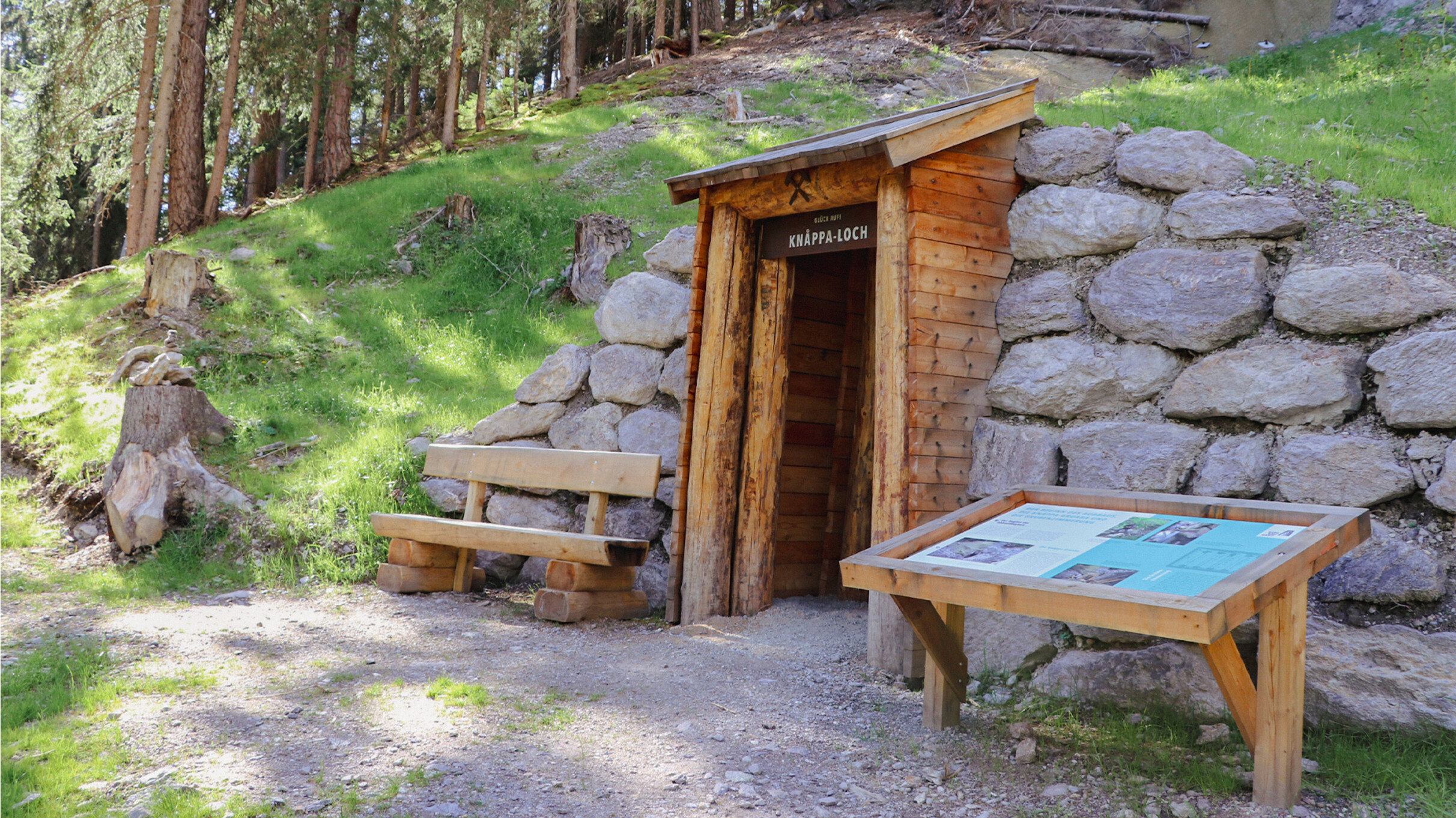 Mobilitätskarte St. Anton - Bergbau Gand Historischer Grubeneingang zum sogenannten „Knappa Loch“ im Wald mit einer Informationstafel und einer Holzbank in der Nähe von St. Anton am Arlberg.