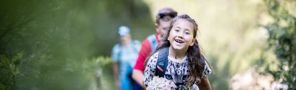 Fröhliches Mädchen mit Blumenstrauß wandert mit Familie durch grüne Klamm bei St. Anton am Arlberg.