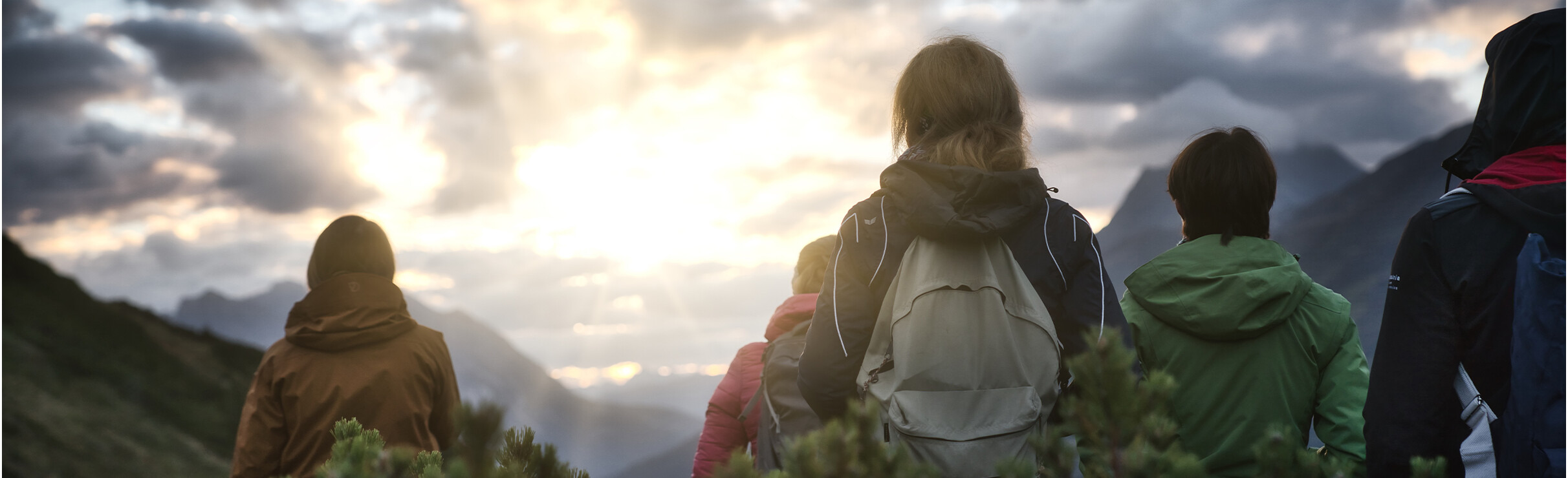Wandergruppe blickt bei Sonnenaufgang über die Berge. Die Szene ist in ruhigem Licht gehalten, die Personen tragen Outdoorjacken und genießen den Blick in die Ferne.