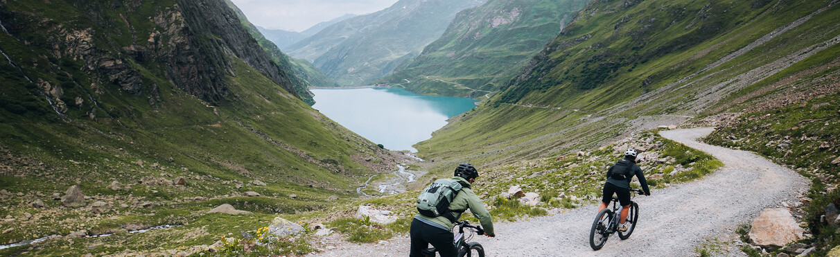 Zwei E-Biker fahren auf einem Schotterweg talwärts mit Blick auf einen türkisblauen Bergsee. Umgeben von steilen, grünen Berghängen.