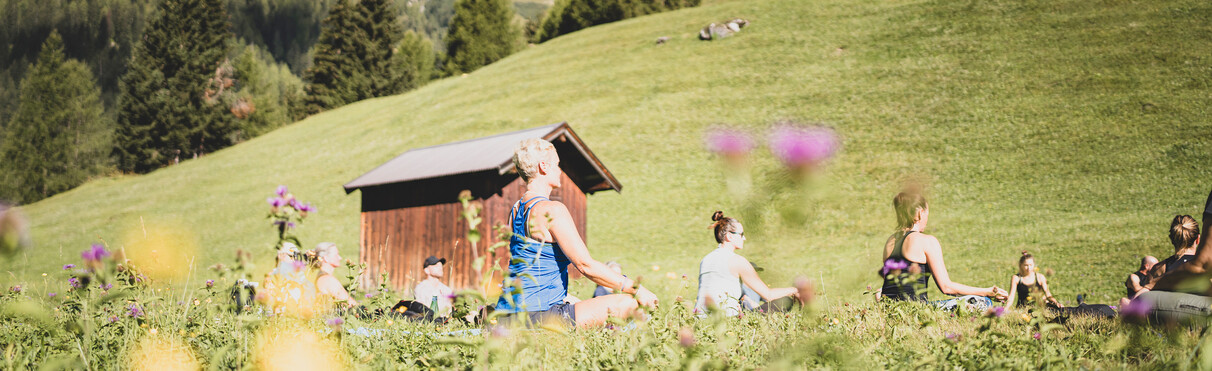 A group practices seated yoga on a sunny alpine meadow with a lush mountain backdrop.