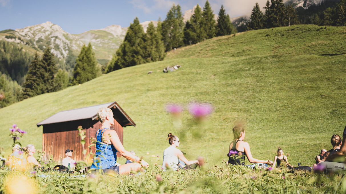 Eine Gruppe praktiziert Yoga im Sitzen auf einer sonnigen Almwiese vor grüner Bergkulisse.