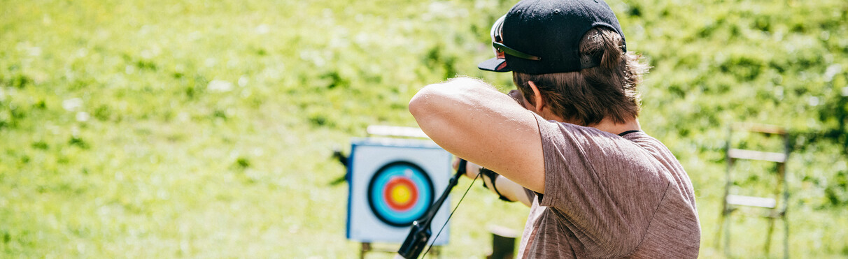 Man in cap aims with bow at a target on a grassy field.