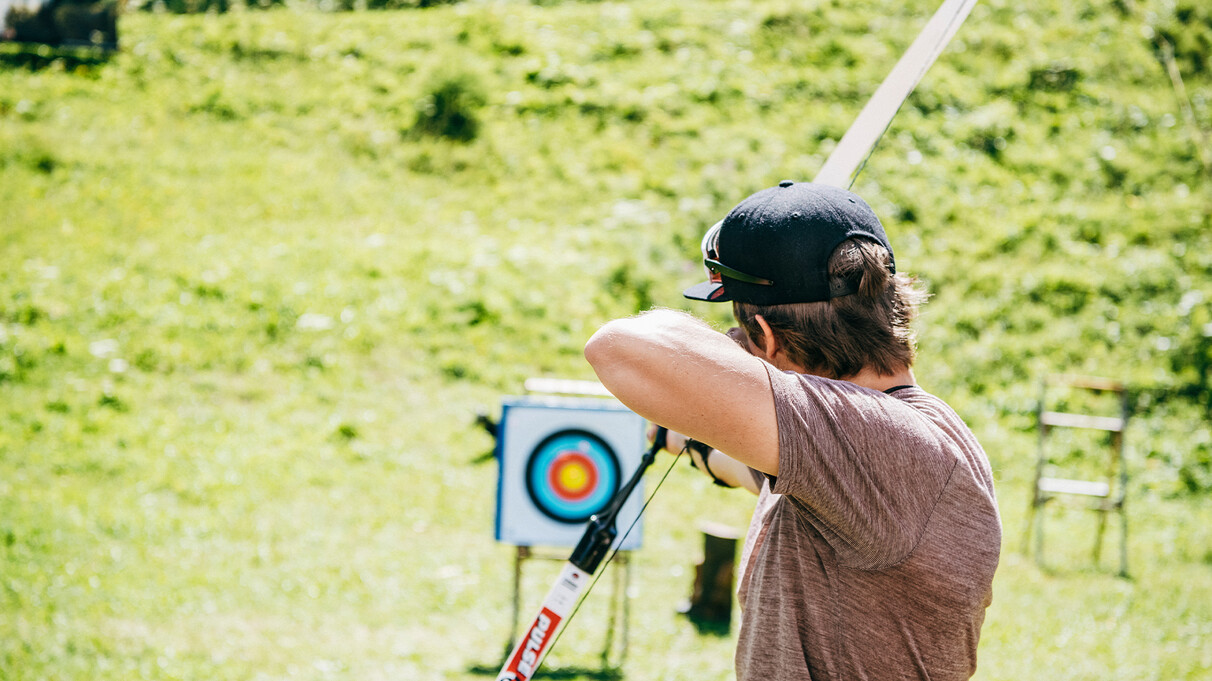Mann mit Kappe zielt beim Bogenschießen auf eine Zielscheibe auf grüner Wiese.