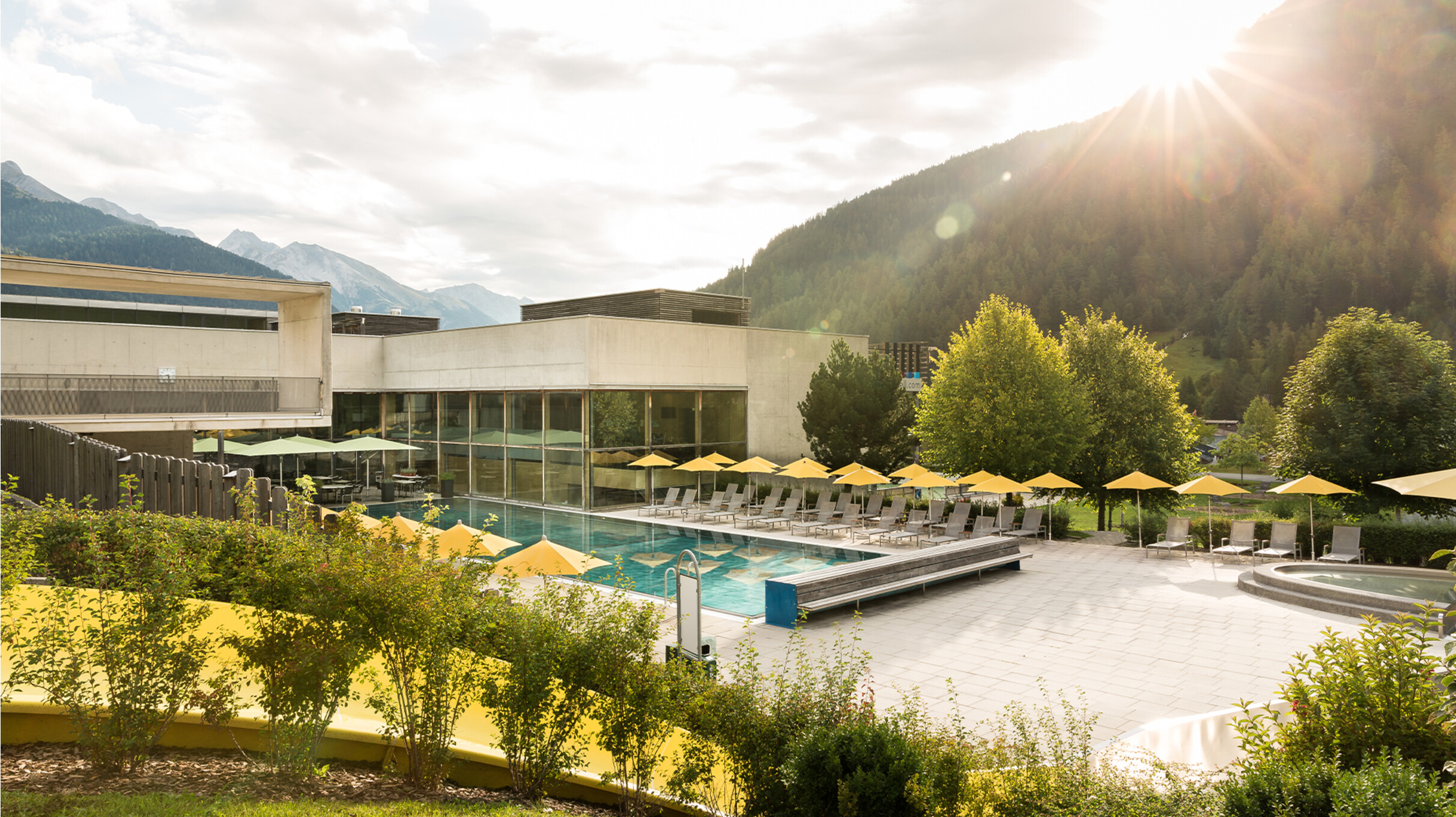 Außenbecken des Arlberg Wellcom (c) DIE WEST Sonnige Poollandschaft mit gelben Schirmen vor moderner Architektur und bewaldeten Berghängen.