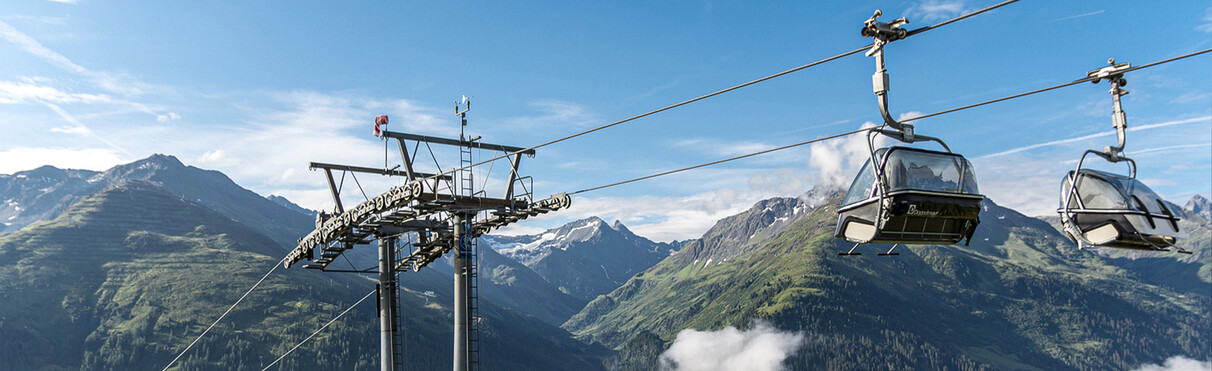 Two empty Gampenbahn chairlifts hover high above a sea of fog with a view of green mountain slopes and peaks near St. Anton am Arlberg.
