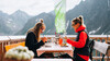 Zwei Personen sitzen auf einer Hüttenterrasse mit Essen vor alpiner Kulisse / Two people sit on a mountain hut terrace eating with alpine peaks in the background