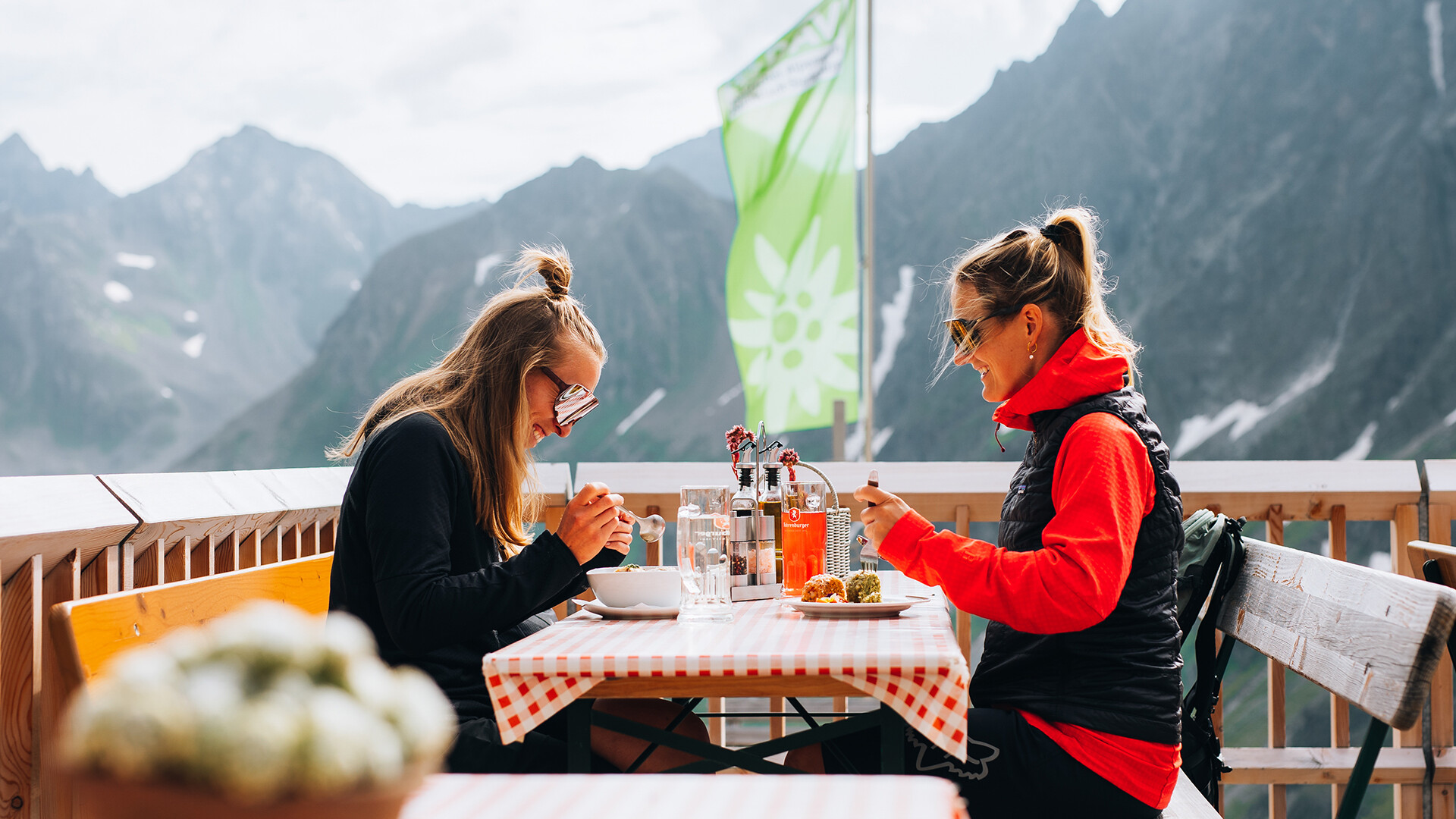 Zwei Personen sitzen auf einer Hüttenterrasse mit Essen vor alpiner Kulisse / Two people sit on a mountain hut terrace eating with alpine peaks in the background