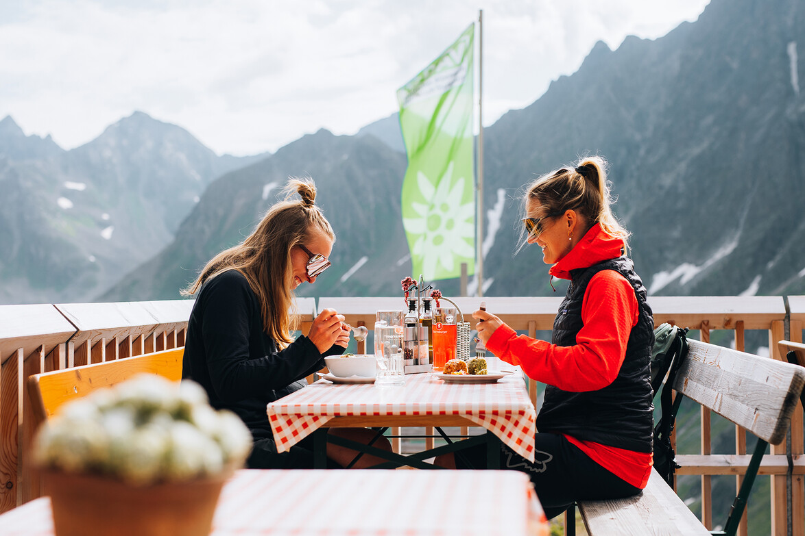 Zwei Personen sitzen auf einer Hüttenterrasse mit Essen vor alpiner Kulisse / Two people sit on a mountain hut terrace eating with alpine peaks in the background
