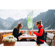 Zwei Personen sitzen auf einer Hüttenterrasse mit Essen vor alpiner Kulisse / Two people sit on a mountain hut terrace eating with alpine peaks in the background