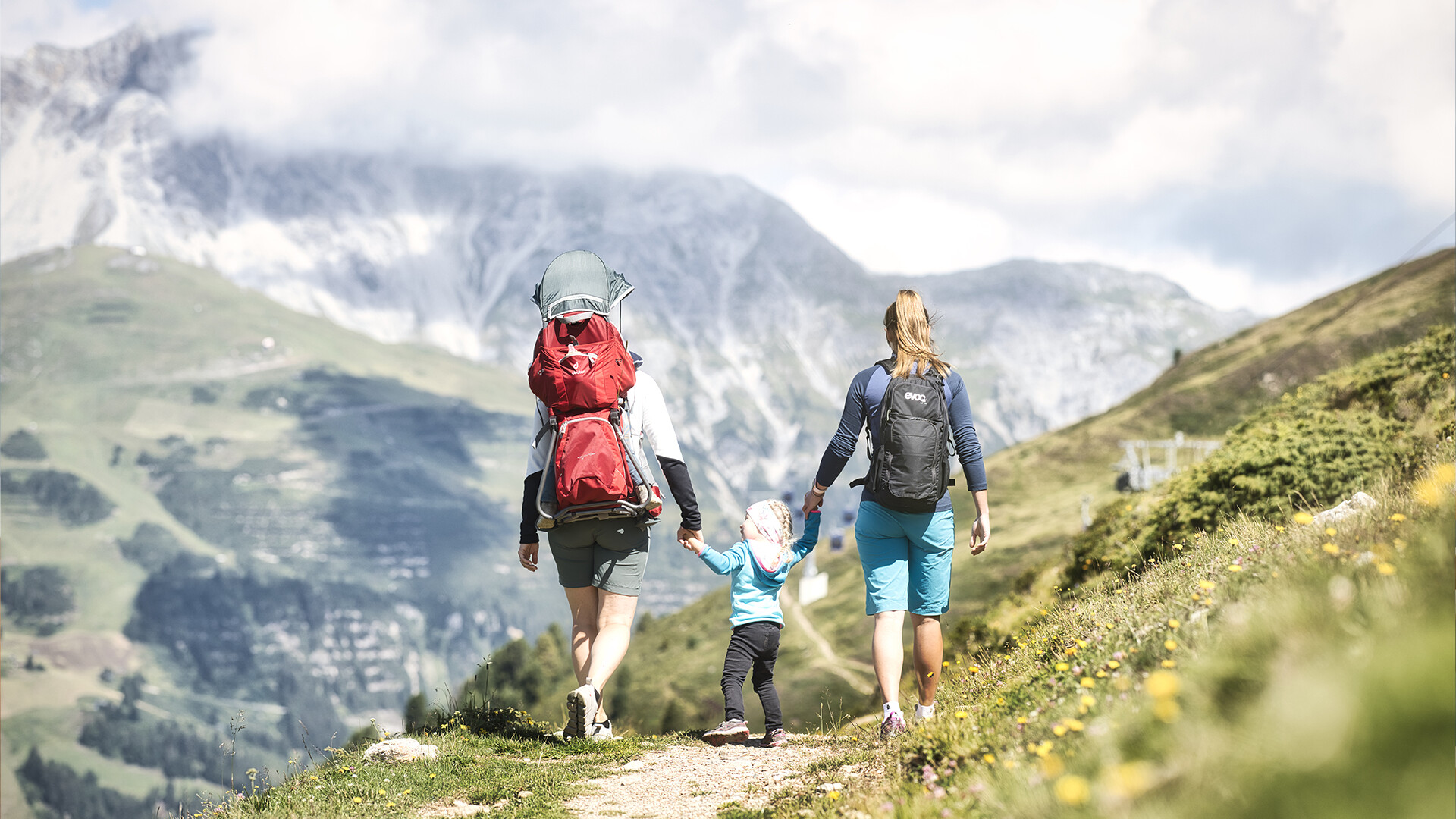 Familie wandert gemeinsam einen alpinen Weg mit blühenden Wiesen und Bergkulisse / Family hikes along an alpine trail with blooming meadows and mountain scenery