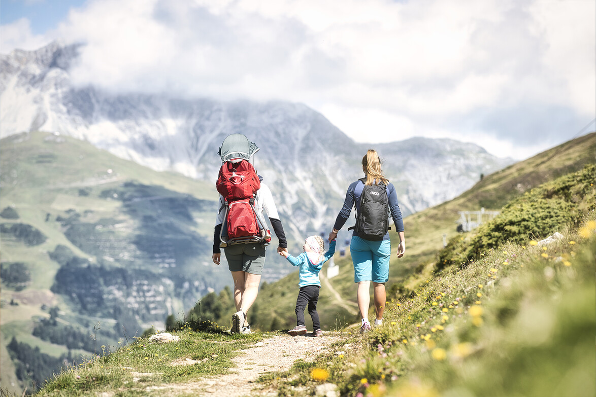 Familie wandert gemeinsam einen alpinen Weg mit blühenden Wiesen und Bergkulisse / Family hikes along an alpine trail with blooming meadows and mountain scenery