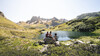 Drei Wanderer rasten an einem kleinen See mit Blick auf die schroffen Berge des Arlbergs / Two hikers rest by a small alpine lake with rugged Arlberg mountains in the background