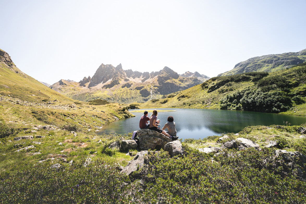 Drei Wanderer rasten an einem kleinen See mit Blick auf die schroffen Berge des Arlbergs / Two hikers rest by a small alpine lake with rugged Arlberg mountains in the background