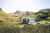 Bergsee-Rast / Alpine Lake Rest Drei Wanderer rasten an einem kleinen See mit Blick auf die schroffen Berge des Arlbergs / Two hikers rest by a small alpine lake with rugged Arlberg mountains in the background