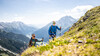 Zwei Wanderer mit Stöcken steigen einen steilen Hang mit Blick ins Tal hinauf / Two hikers with poles ascend a steep slope with a wide view into the valley