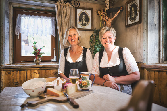 Zwei Damen in traditioneller Tiroler Tracht sitzen am Esstisch und auf der "Eckbank" vor ihnen Zutaten für die Flirscher Kaskiachla