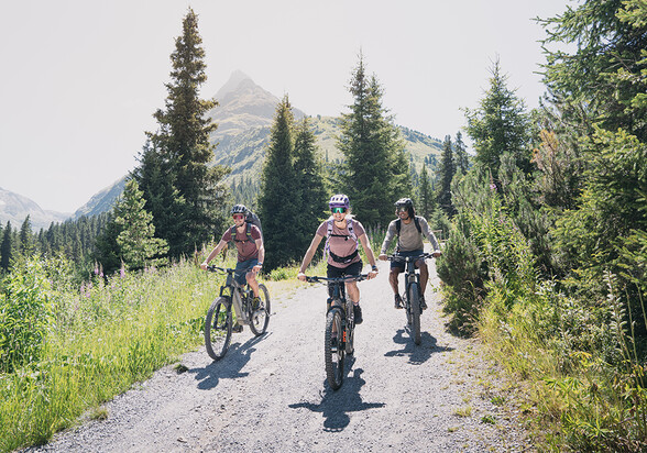 Drei Radfahrer:innen fahren auf einem sonnigen Schotterweg durch den grünen Bergwald in St. Anton am Arlberg.