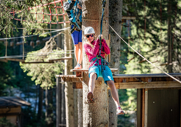 Frau mit Helm saust lachend an einer Seilrutsche durch den Hochseilgarten im Wald bei St. Anton.