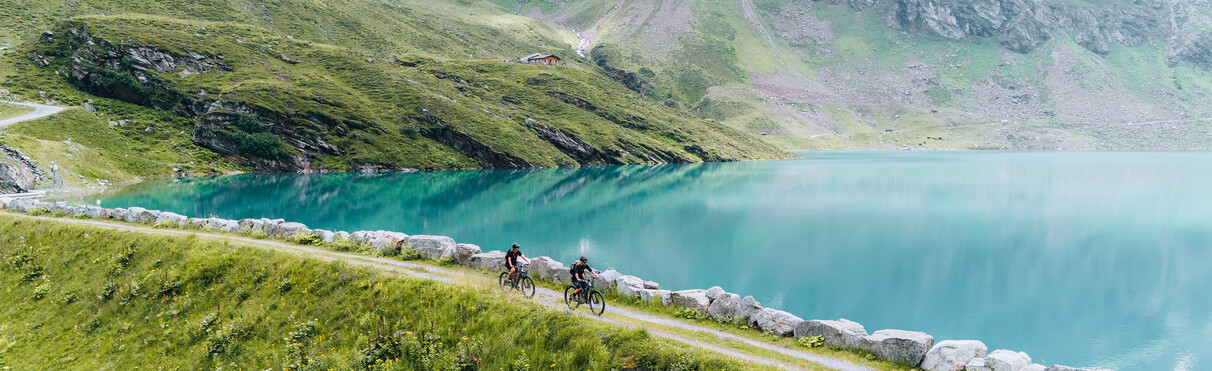 Zwei Mountainbiker fahren entlang eines türkisblauen Bergsees mit hochalpiner Landschaft im Hintergrund.