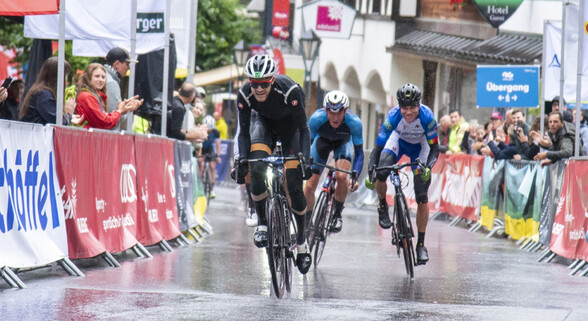 Die drei Führenden beim Arlberg Giro beim Zieleinlauf in der verregneten Fußgängerzone von St. Anton am Arlberg mit jubelnden Fans hinter den Absperrungen.