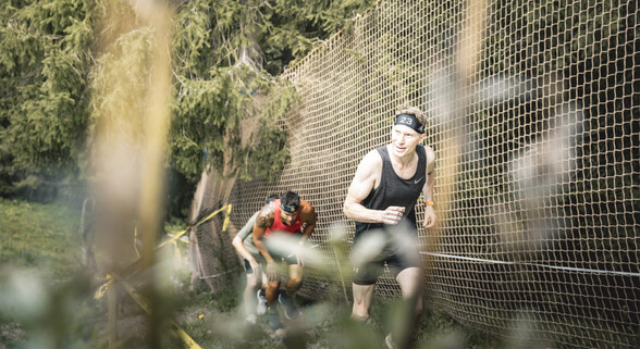 Blick herab auf drei männliche Teilnehmer des Arlberger Wadlbeisser die gerade einen steilen Hang auf einem Trampelpfad bezwingen . Neben dem Hang sind dichte Bäume und eine Absperrung.
