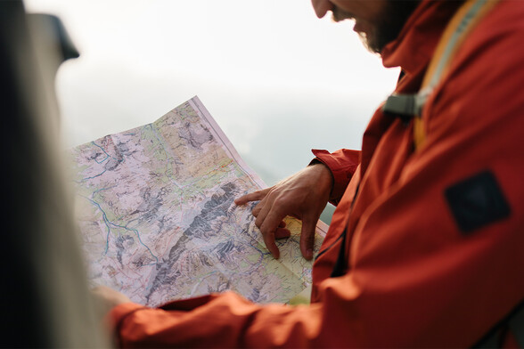 Ein Mann in oranger Wanderjacke hält eine Wanderkarte in der Hand und zeigt auf eine Route. Im Hintergrund ist die Landschaft unscharf und lichtdurchflutet.