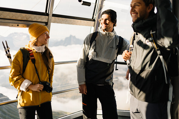 Drei Personen in Wanderkleidung stehen in einer Gondel. Sie lächeln einander an, im Hintergrund sind Berge und Wolken zu sehen. Die Szene wirkt freundlich und voller Vorfreude.