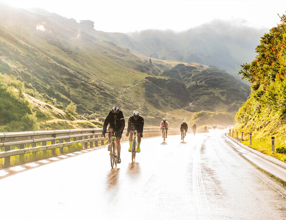 Radfahrer fahren bei Sonnenaufgang auf einer nassen Bergstraße. Die Landschaft ist von Licht und Nebel umgeben.