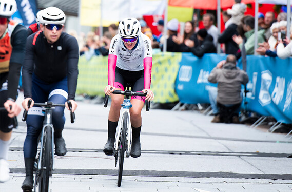 Mehrere Radfahrer erreichen erschöpft das Ziel, während die Menge sie anfeuert. Eine Fahrerin in weiß-rosa Trikot fährt vorne.